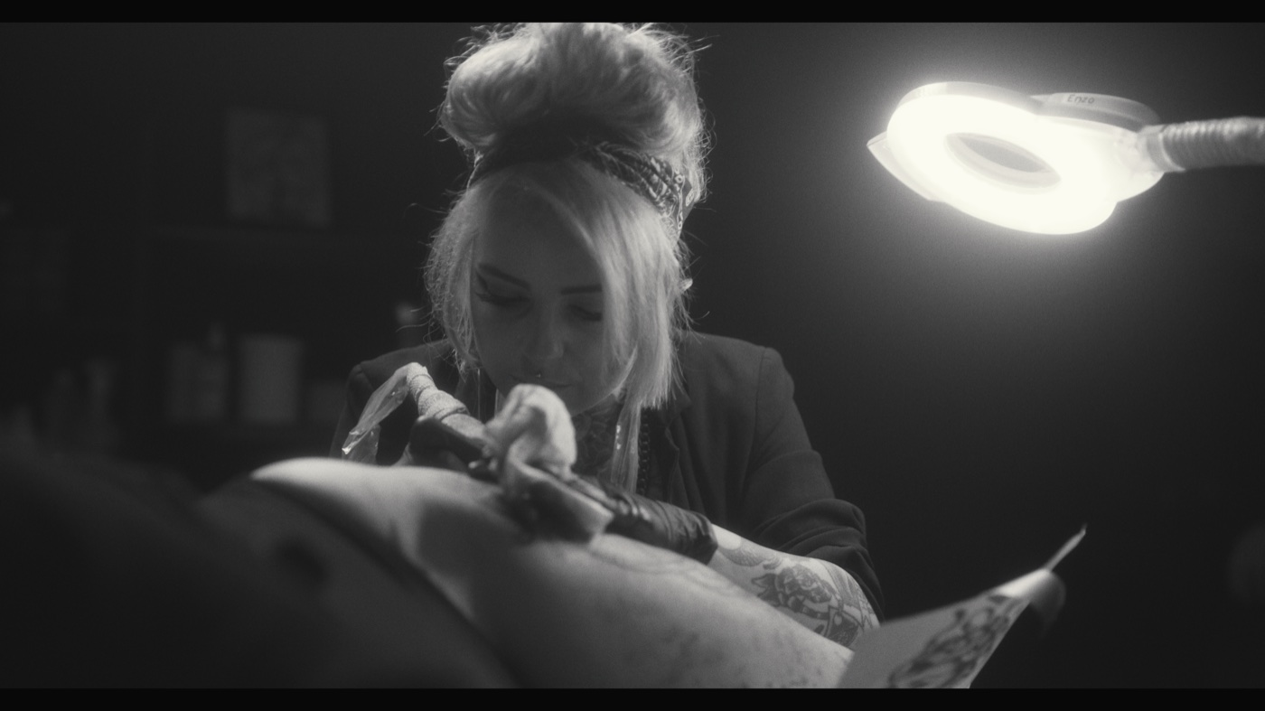 A tattoo artist working on a client beneath a bright circular task light in black and white.