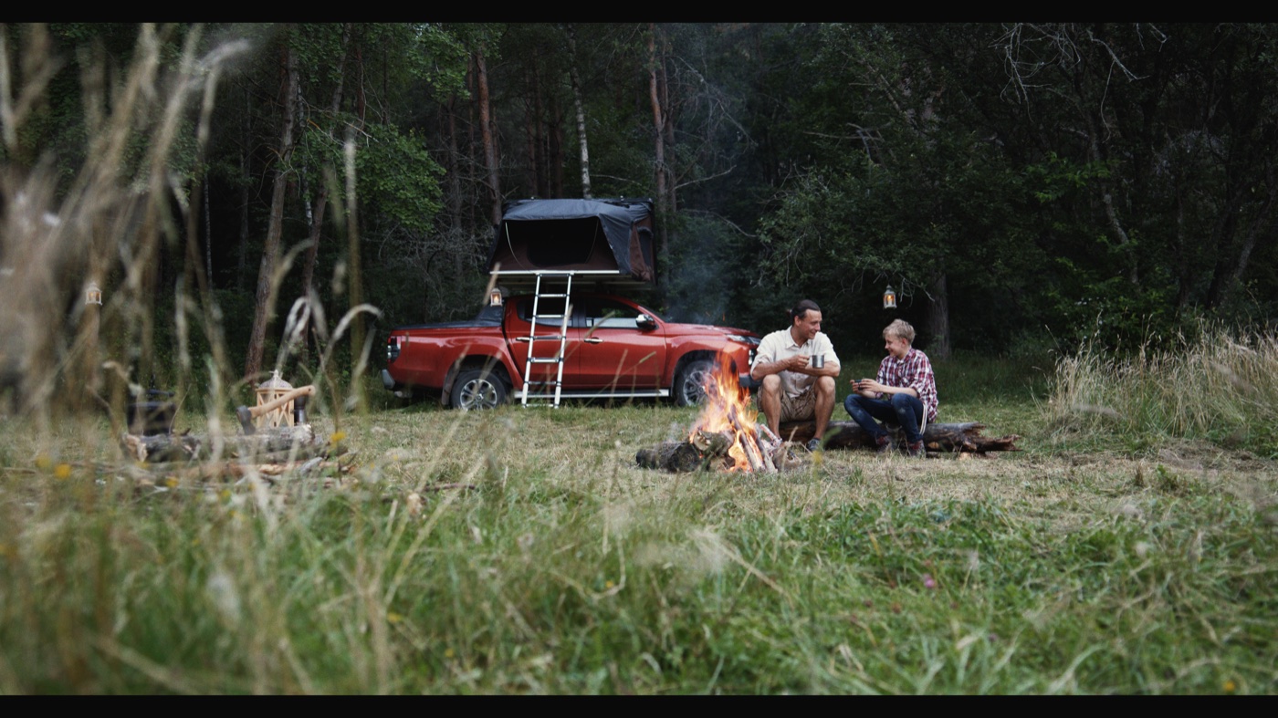 Two people sitting beside a campfire near a red pickup with a rooftop tent in a forest clearing.
