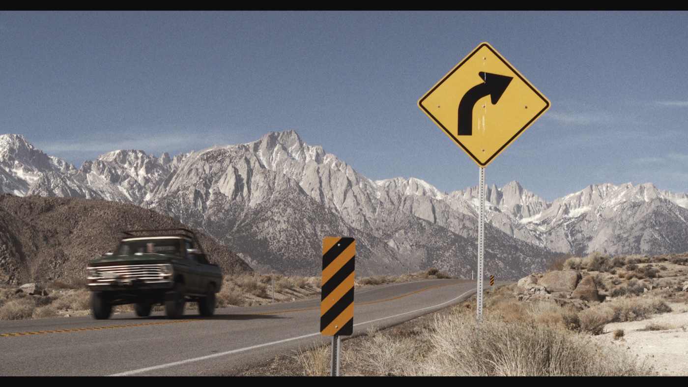 A dark pickup passing a yellow curve sign on a desert road beneath pale mountains and a blue sky.