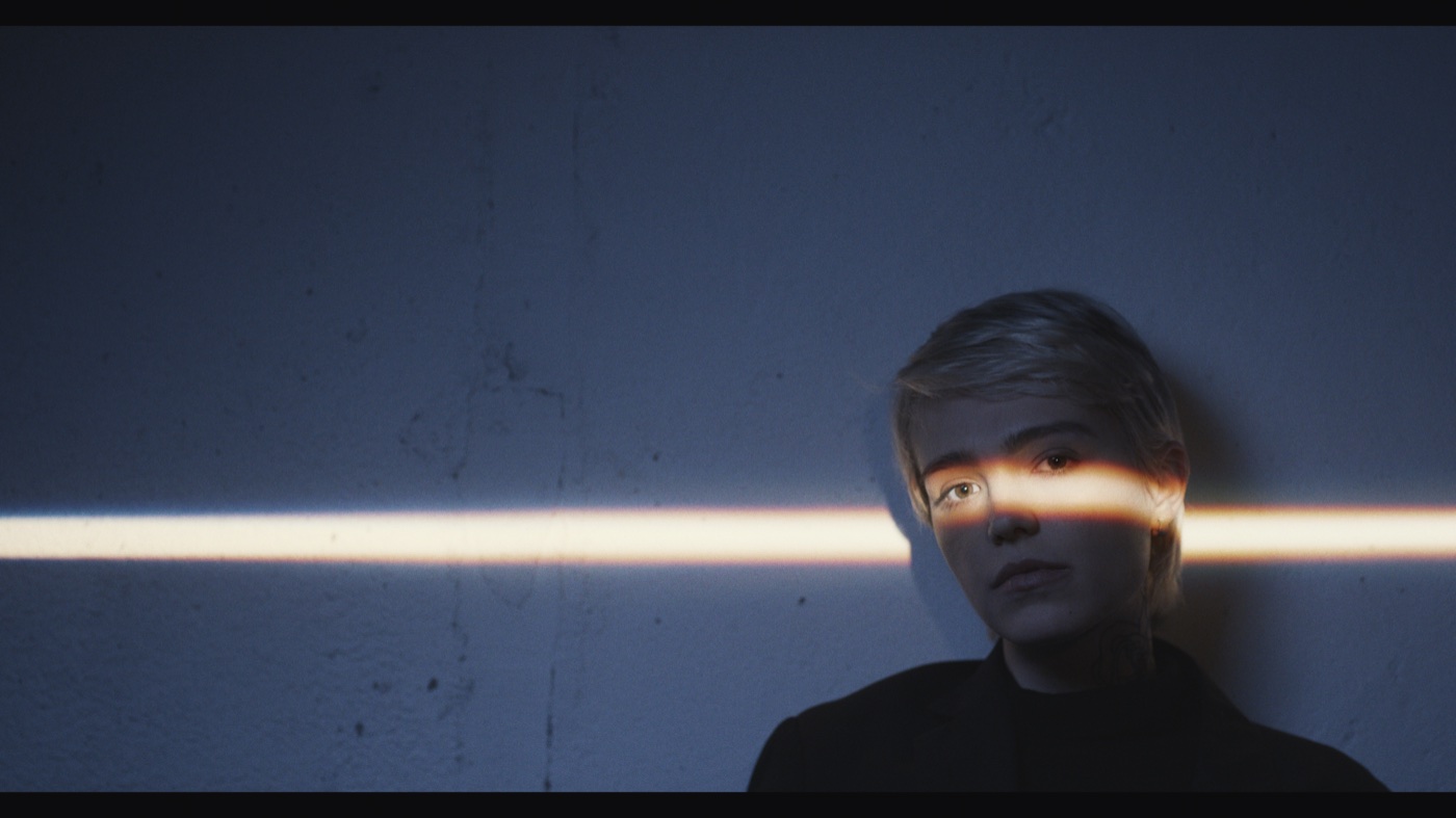 A short-haired person against a blue wall with a narrow band of warm light crossing their eyes.