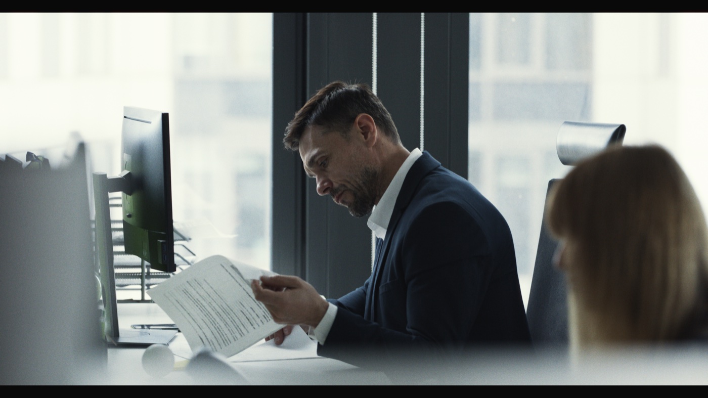 A man reading paperwork at an office desk in front of large, brightly lit windows.